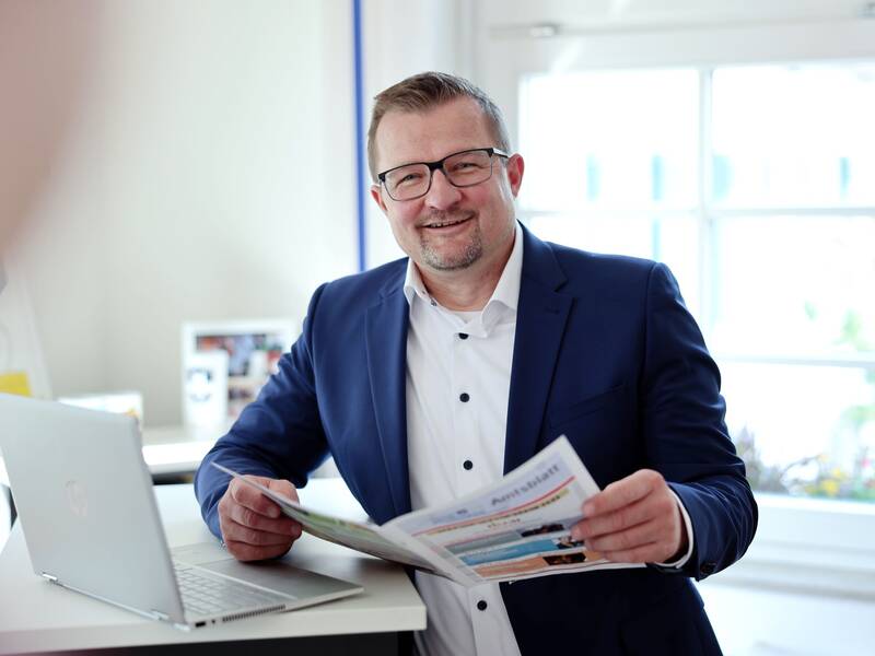 Bürgermeister Stefan Wörner steht lächelnd in einem blauen Anzug in seinem Büro neben einem Laptop und hält das Amtsblatt in den Händen.