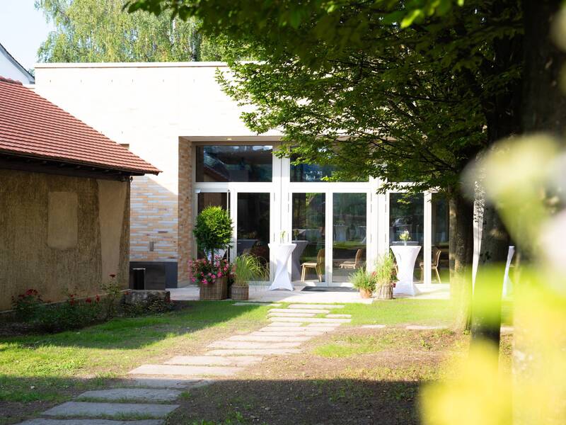 Ein gepflegter Gartenweg führt zu einem modernen Gebäude, dem Kulturhaus Klosterkirche, mit großen Fenstern, flankiert von dekorativen Pflanzen und Stehtischen.