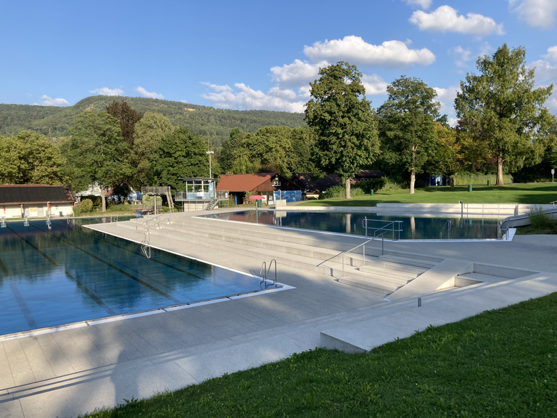 Ein großzügiges Freibad mit Treppen­eingang, umgeben von Bäumen und einer Wiese, vor einer hügeligen Landschaft.